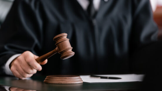 This photo shows a partial view of a blurred judge in robe holding gavel in court