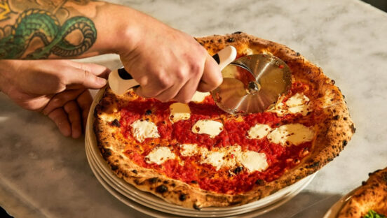 This photo shows a man's tattooed arm as he slices a Margherita pizza with a beautifully blistered crust.