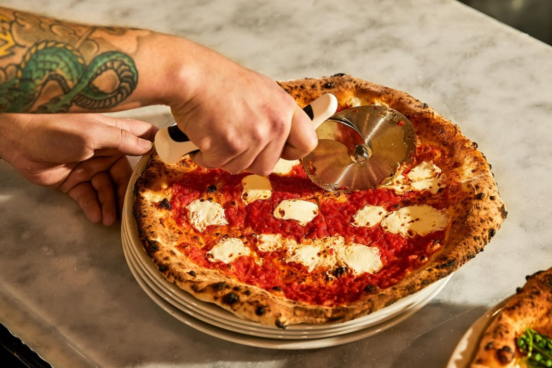 This photo shows a man's tattooed arm as he slices a Margherita pizza with a beautifully blistered crust.