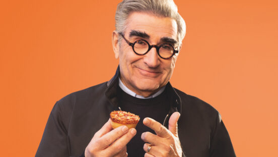 This photo shows Eugene Levy, wearing glasses and a dark shirt, grinning and holding a Little Caesars Crazy Puff.