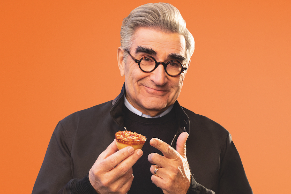 This photo shows Eugene Levy, wearing glasses and a dark shirt, grinning and holding a Little Caesars Crazy Puff.