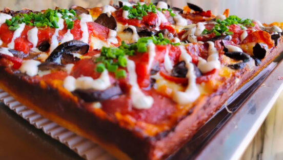 This photo shows a Detroit-style pizza topped with pepperoni, bacon, mushrooms, a ranch drizzle and fresh chives.