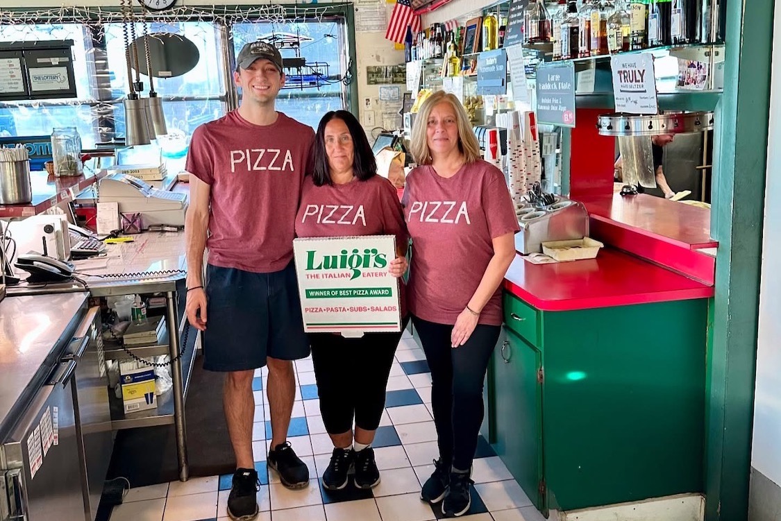 This photo shows Justin Pioppi with his mother and another woman standing in Luigi's Pizzeria. His mom, standing in the middle, is holding a branded Luigi's pizza box. All three are wearing red t-shirts with the word "Pizza."