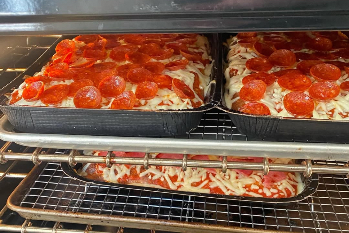 This photo shows two pepperoni pizzas baking in a pan on an oven rack. Beneath them is another pizza with mozzarella and Canadian bacon.