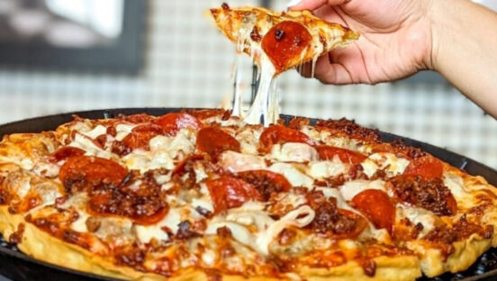 This photo shows a person's hand removing a slice from a whole thin-crust pizza with a nice cheese pull.