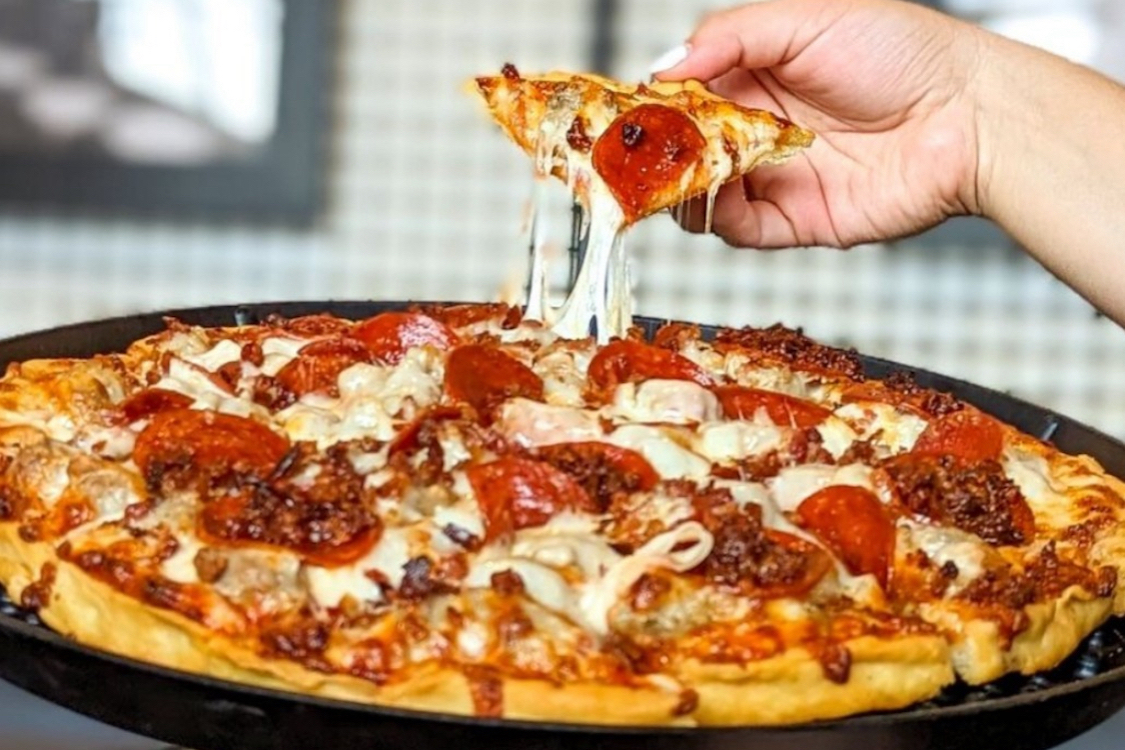 This photo shows a person's hand removing a slice from a whole thin-crust pizza with a nice cheese pull.