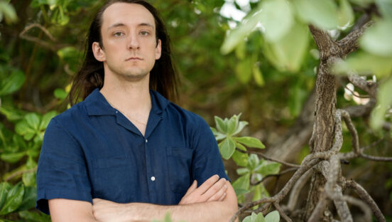This photo shows Justin Pioppi, wearing a blue shirt, arms folded and looking serious, on the island of Fiji.