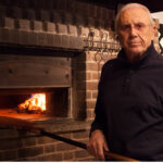 This photo shows a black-clad Patsy Grimaldi holding a long peel and standing in front of a coal-fired oven.