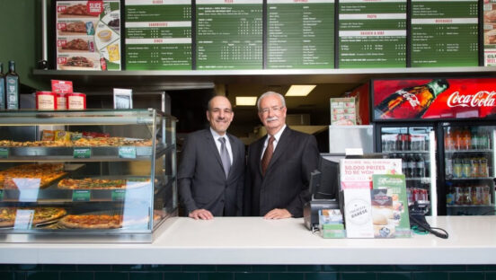 This photo shows the Pizza Nova founder and his son, the current president, both wearing suits and standing side by side behind the ordering counter of one of the restaurants.