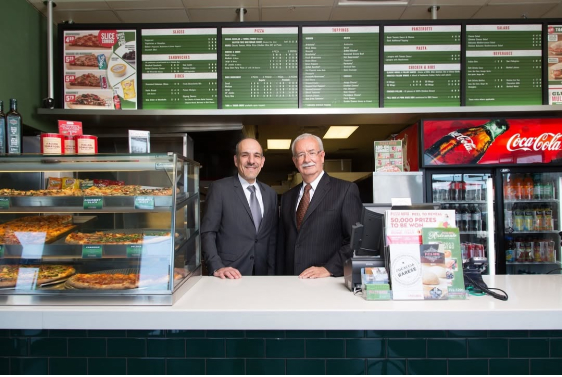 This photo shows the Pizza Nova founder and his son, the current president, both wearing suits and standing side by side behind the ordering counter of one of the restaurants.