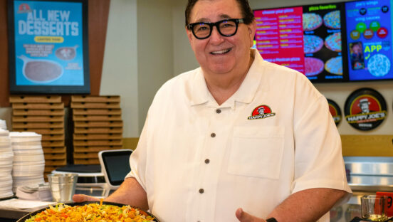This photo shows Happy Joe's CEO Tom Sacco wearing a white shirt and posing with a taco pizza.