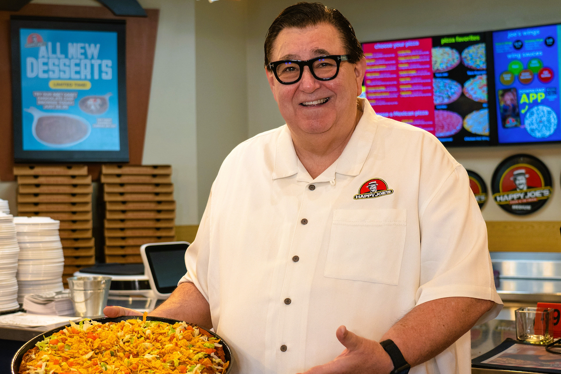 This photo shows Happy Joe's CEO Tom Sacco wearing a white shirt and posing with a taco pizza.