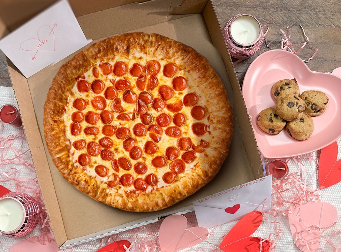 This photo shows an overhead view of Vocelli's Stuffed Crust pizza, topped with pepperoni, in a delivery box surrounded by paper hearts, pink string and an order of chocolate chip cookies in a little heart-shaped box.