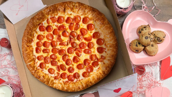 This photo shows an overhead view of Vocelli's Stuffed Crust pizza, topped with pepperoni, in a delivery box surrounded by paper hearts, pink string and an order of chocolate chip cookies in a little heart-shaped box.