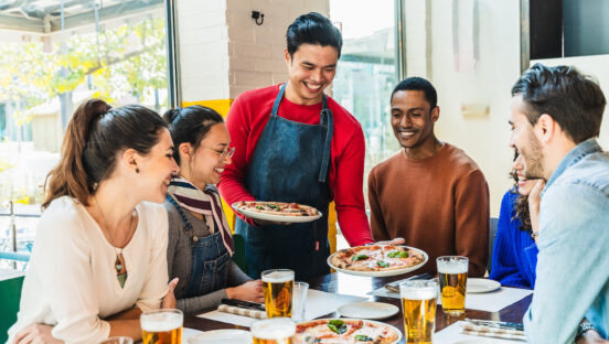 This photo shows a smiling young waiter serving pizza to a diverse group of customers.