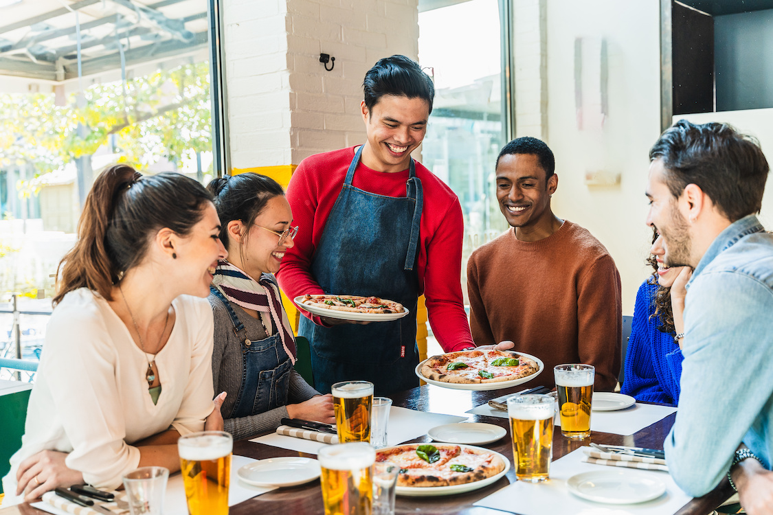 This photo shows a smiling young waiter serving pizza to a diverse group of customers.