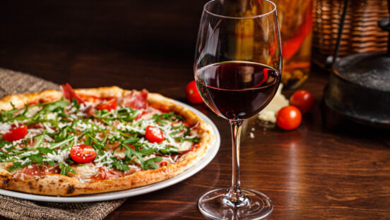 This photo shows a glass of red wine next to a thin-crust pizza with prosciutto, cherry tomatoes, arugula and Parmesan
