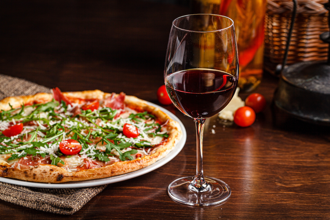 This photo shows a glass of red wine next to a thin-crust pizza with prosciutto, cherry tomatoes, arugula and Parmesan