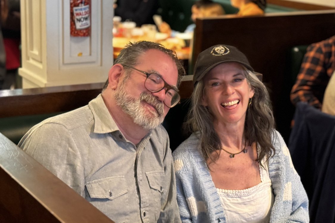 This photo shows Andrea, wearing a cap, sitting in a booth next to her bearded twin brother.