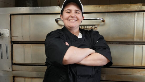 This photo shows Victoria Tiso, wearing a black shirt, standing in front of her deck oven with her arms crossed and a proud smile.