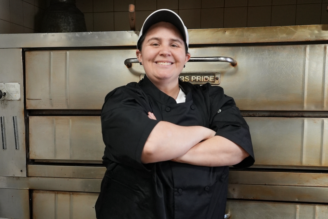 This photo shows Victoria Tiso, wearing a black shirt, standing in front of her deck oven with her arms crossed and a proud smile.