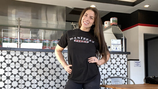This photo shows Christina Martin, dressed in a black t-shirt and black jeans, standing with her hands on her hips and smiling in Manizza's Pizza.