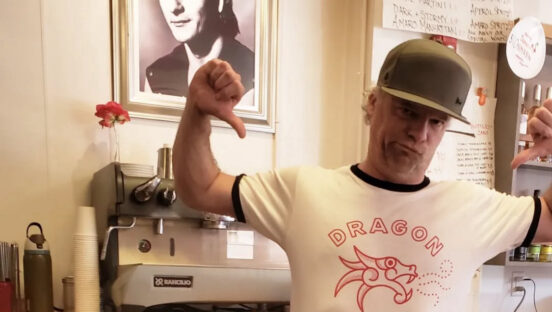 This photo shows Charlie Redd pointing with his thumbs at his Dragon Pizza-branded t-shirt while standing in front of a framed photo of Patrick Swayze.