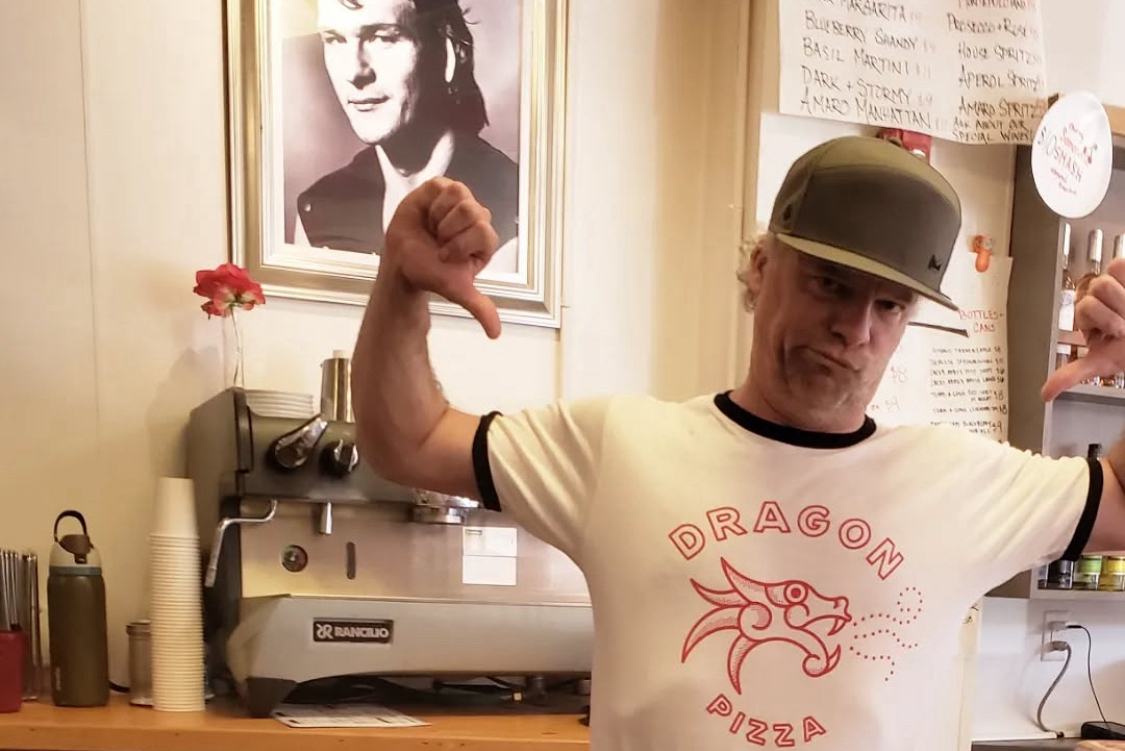 This photo shows Charlie Redd pointing with his thumbs at his Dragon Pizza-branded t-shirt while standing in front of a framed photo of Patrick Swayze.