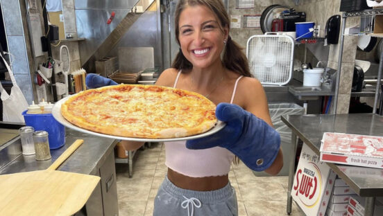 This photo shows a smiling Giuliana Caliscibetta holding up a large cheese pizza in a pan.