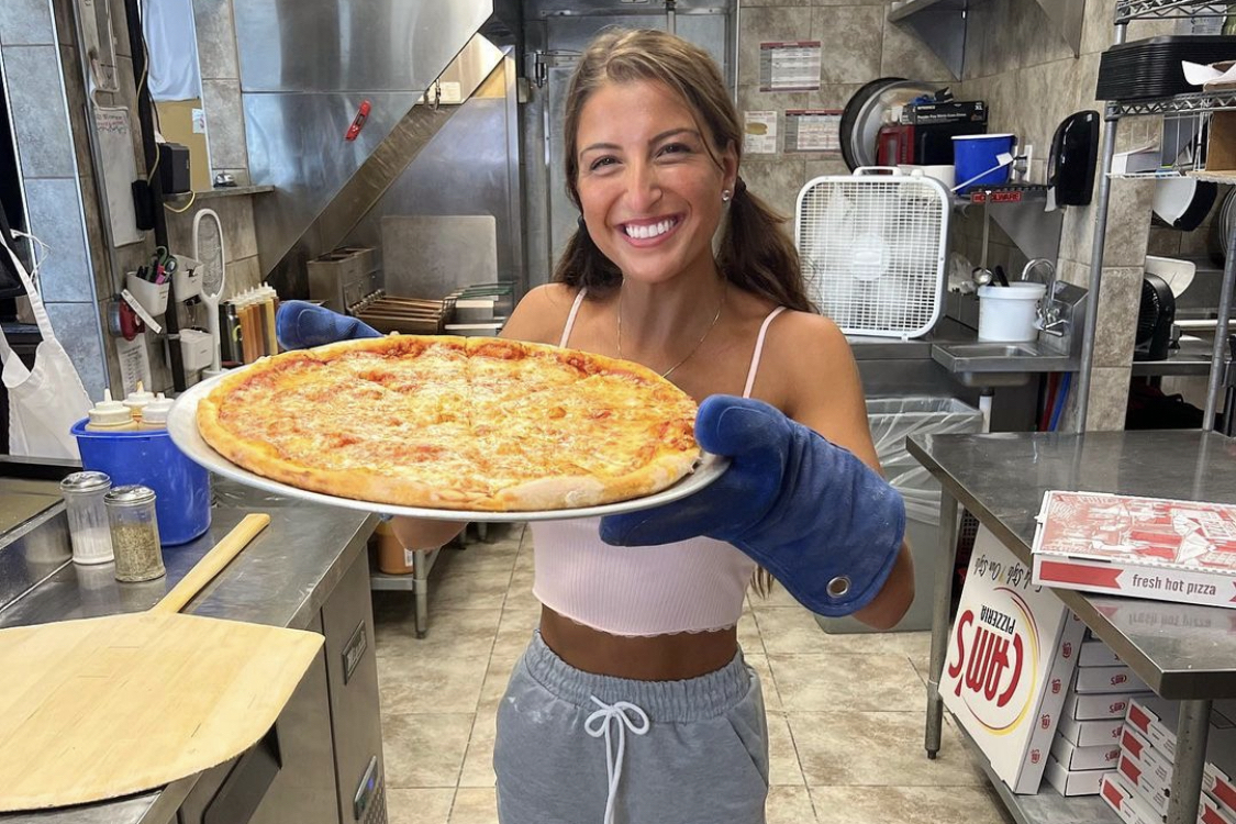 This photo shows a smiling Giuliana Caliscibetta holding up a large cheese pizza in a pan.