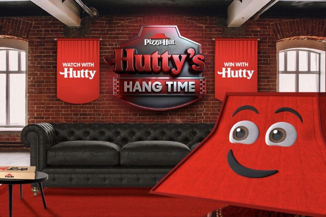 This is an illustration of Hutty in a living room or den, featuring a sort of lampshade (red in color) with eyes and a big smile in the foreground representing Hutty and, in the background, a black sofa, a red-brick wall, banners that read Win With Hutty and Watch With Nutty and, between them, a sign that reads Pizza Hut Hutty's Hang Time. Near the sofa is a table with a Pizza Hut box.
