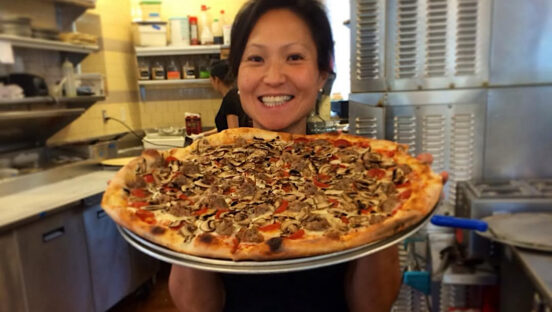 This photo shows a smiling Ann Kim holding a platter with a large pizza in the kitchen of one of her restaurants.