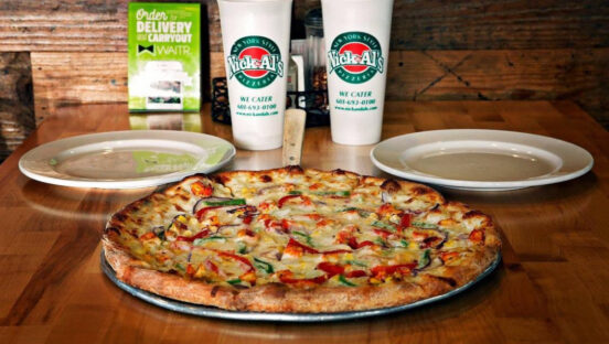 This photo shows a pizza on a tray, flanked by plates, and branded Nick & Al's beverage cups behind it.
