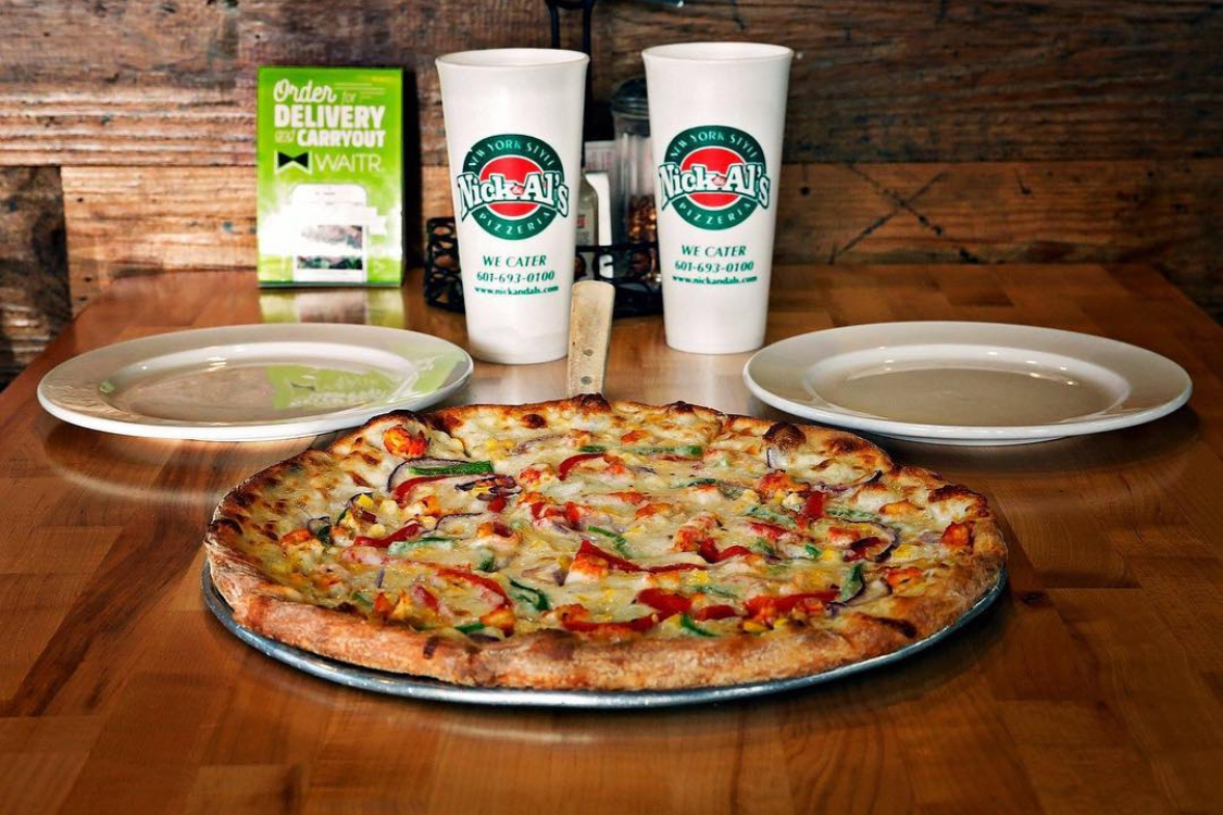 This photo shows a pizza on a tray, flanked by plates, and branded Nick & Al's beverage cups behind it.