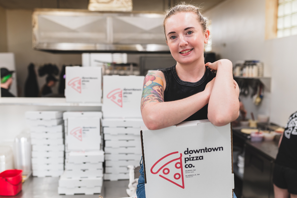 This photo shows a smiling Lizz Aquarian, with a prominent pizza tattoo on her upper right arm, with a Downtown Pizza Company carryout box.