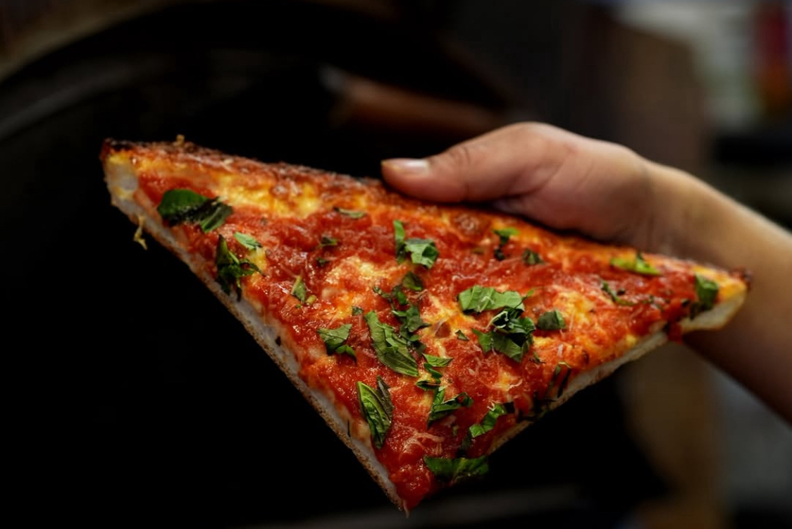 This photo shows a man's hand holding a slice of the Skinny Deep Dish pizza with cheese, sauce and shredded basil.