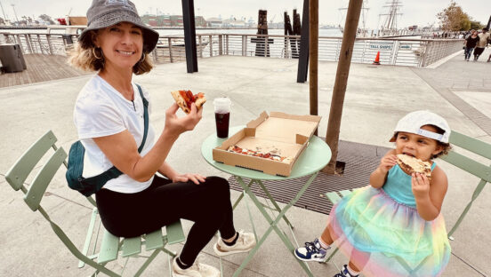 This photo shows Jillana with her daughter, Sofia, who is wearing a cute rainbow-colored skirt. They are both seated at an outdoor table and eating pizza.