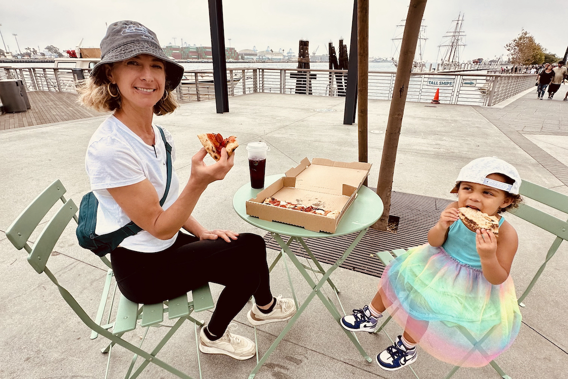 This photo shows Jillana with her daughter, Sofia, who is wearing a cute rainbow-colored skirt. They are both seated at an outdoor table and eating pizza.