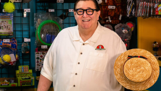 This photo shows Tom Sacco in a Happy Joe's restaurant, wearing a white shirt, smiling and holding a straw hat.