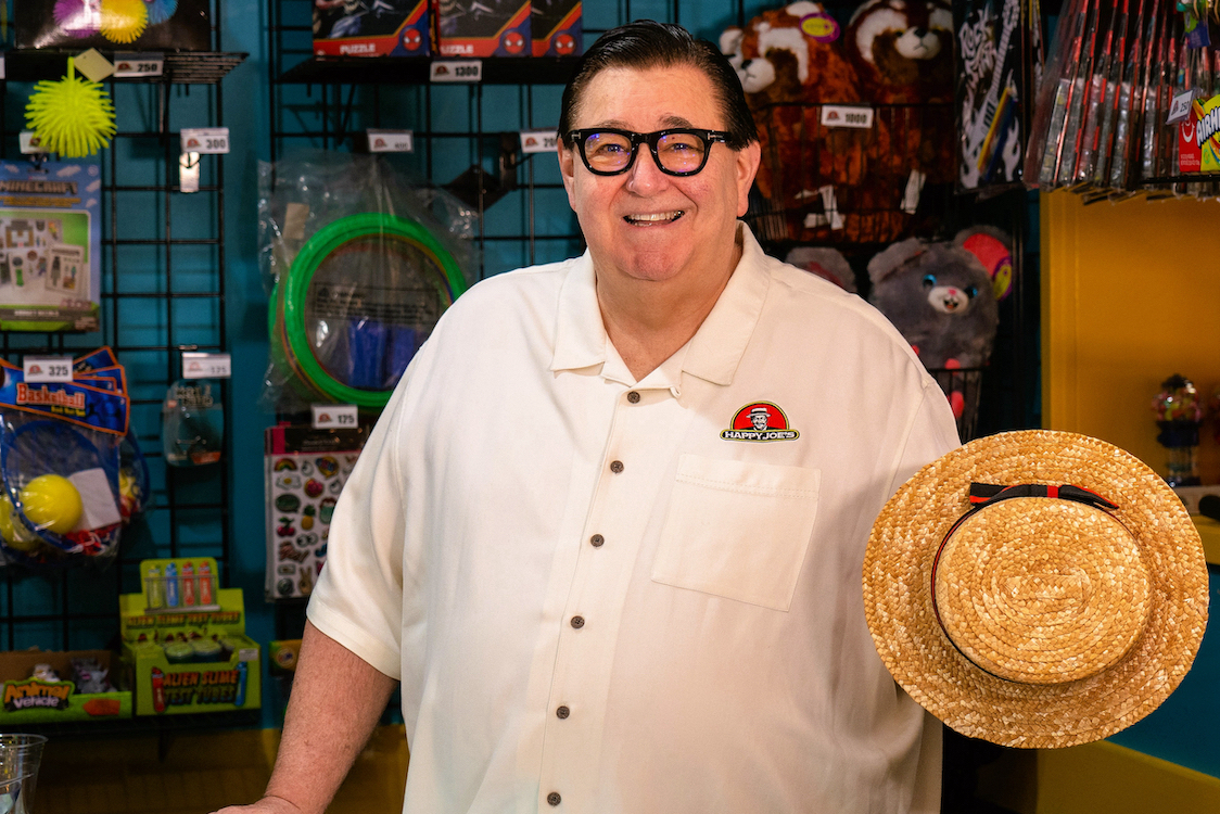 This photo shows Tom Sacco in a Happy Joe's restaurant, wearing a white shirt, smiling and holding a straw hat.