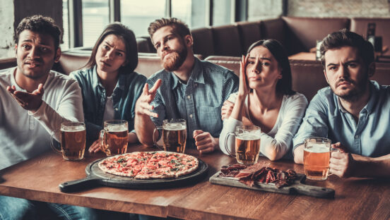 This photo shows a group of five friend eating pizza in a restaurant and watching sports on TV.