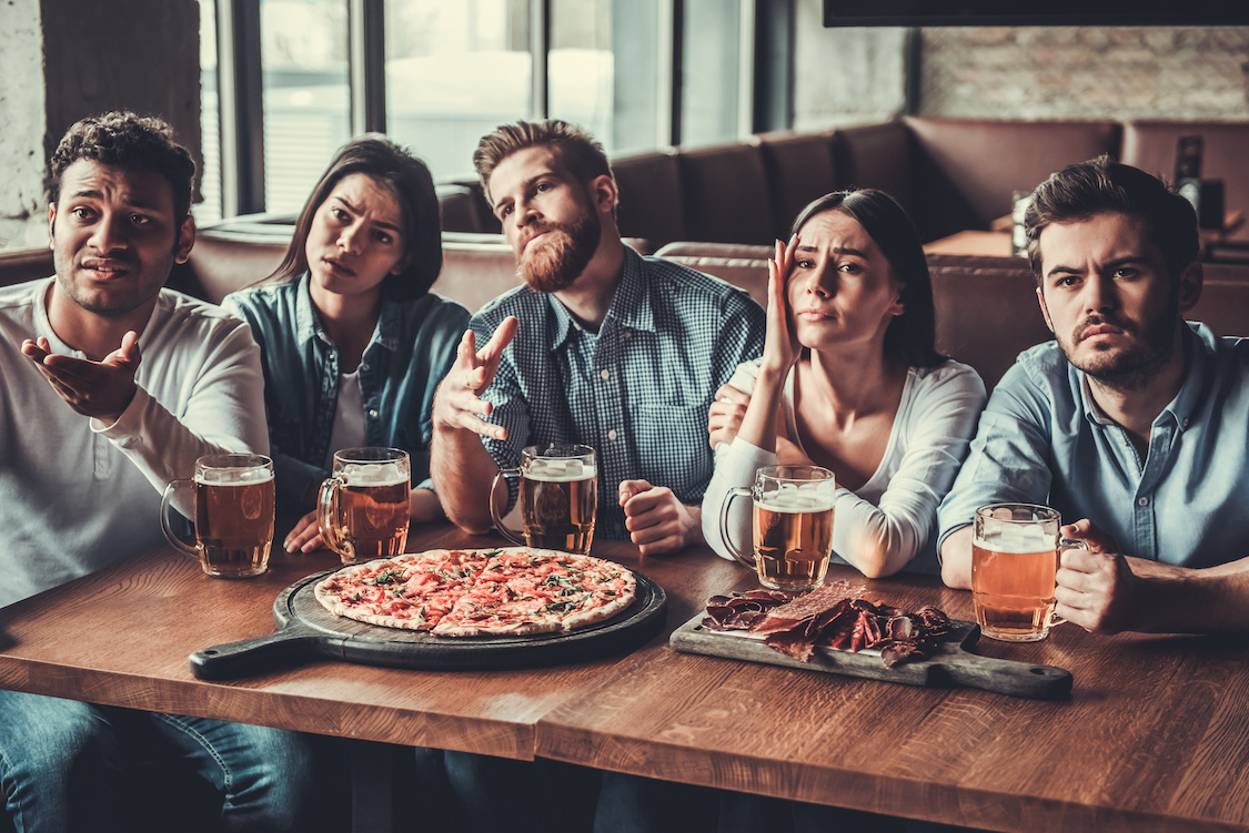 This photo shows a group of five friend eating pizza in a restaurant and watching sports on TV.