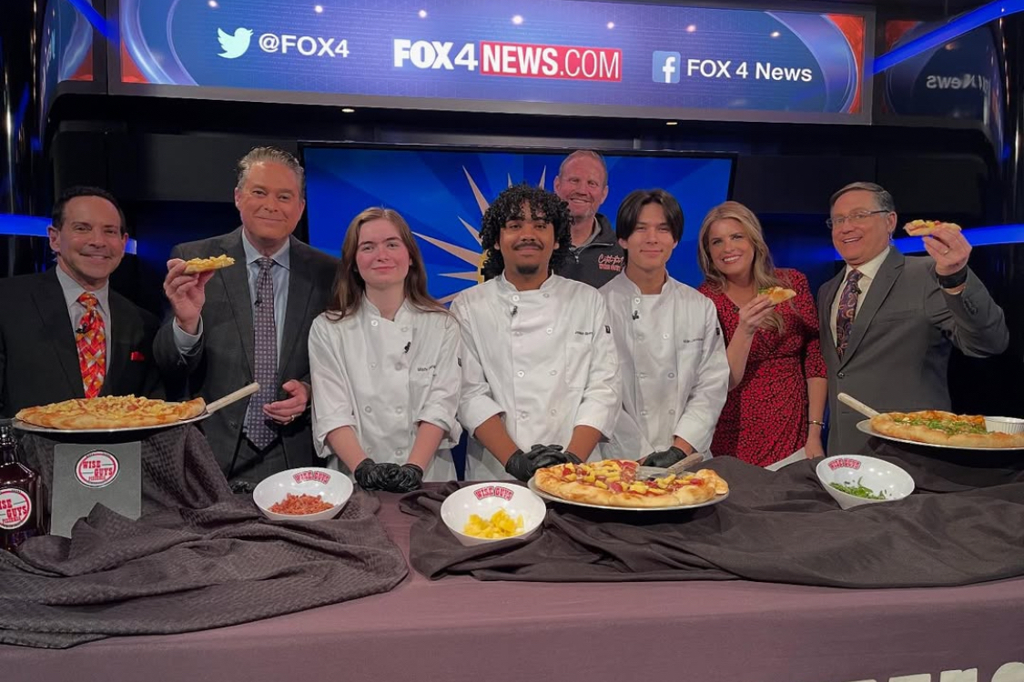This photo shows students from Grapevine-Colleyville Independent School District, wearing white chef coats, posing with the hosts of a Fox 4 TV news show. The TV hosts are holding up slices of pizza.