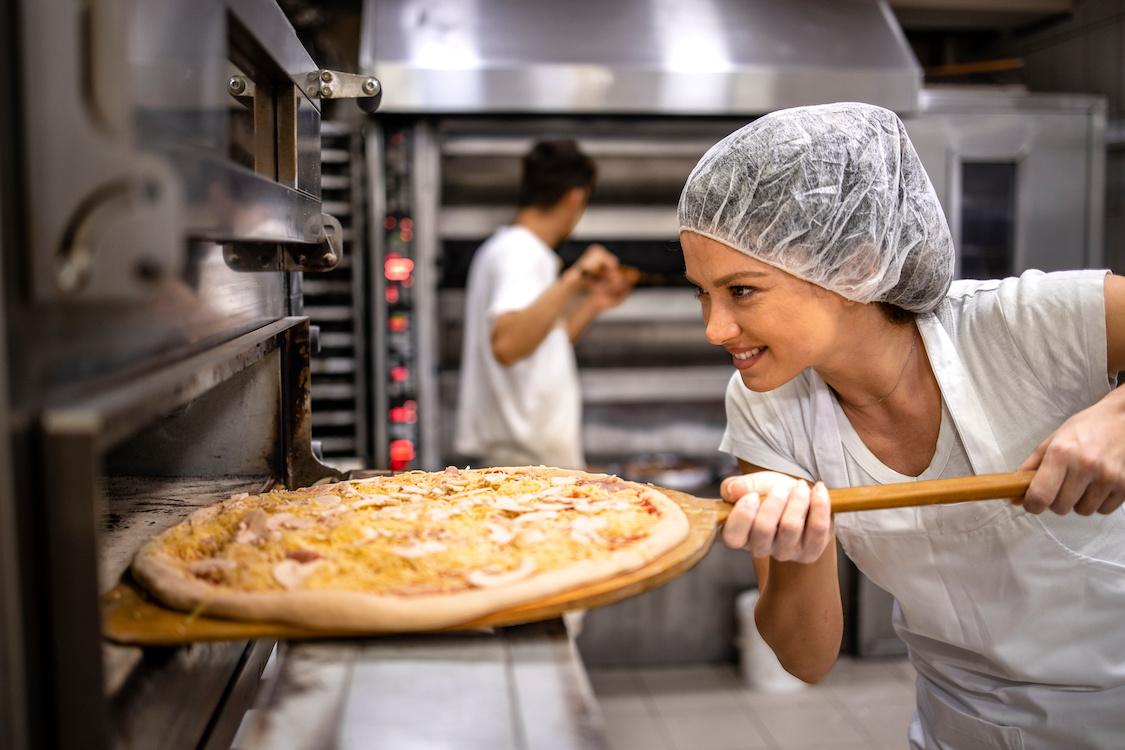 This photo shows a female chef, wearing a white uniform and hair net, putting a pizza into an oven in a restaurant kitchen.