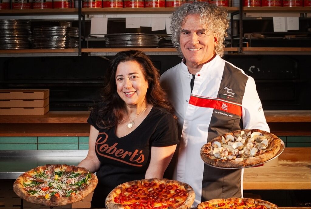 This photo shows Dana Hokin and Robert Garvey holding pizzas and smiling in their dining room at Robert's Pizza Co.
