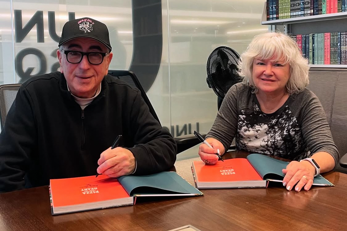 This photo shows Paulie Gee and Mary Ann sitting at a bookstore table signing copies of their new cookbook.
