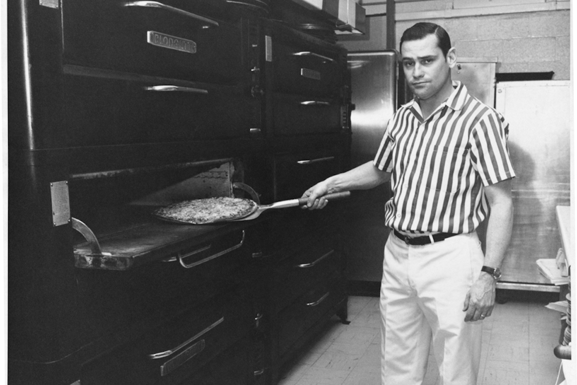 This black and white photo shows Joe Aurelio Jr. peeling a pizza out of an oven in the early 1960s.