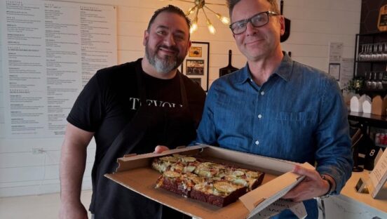 This photo shows Giovanni Labbate in a black t-shirt. He's standing next to Steve Dolinsky in a blue button-down shirt, and the latter is holding a Detroit-style pizza topped with zucchini slices and other ingredients.
