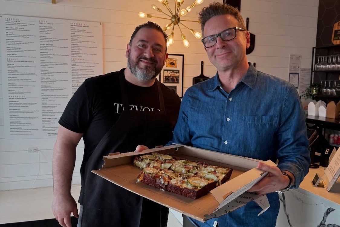 This photo shows Giovanni Labbate in a black t-shirt. He's standing next to Steve Dolinsky in a blue button-down shirt, and the latter is holding a Detroit-style pizza topped with zucchini slices and other ingredients.