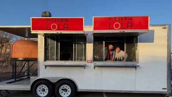 This photo shows two men poking their heads out of one of the windows in the Pizzeria Lola food trailer, with two Pizzeria Lola signs on top of the trailer and a dome-shaped wood fired oven visible on the far left.